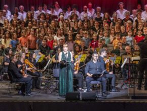 Kammerchor auf einer großen Bühne, Frau in grünem glitzernden Kleid im Vordergrund